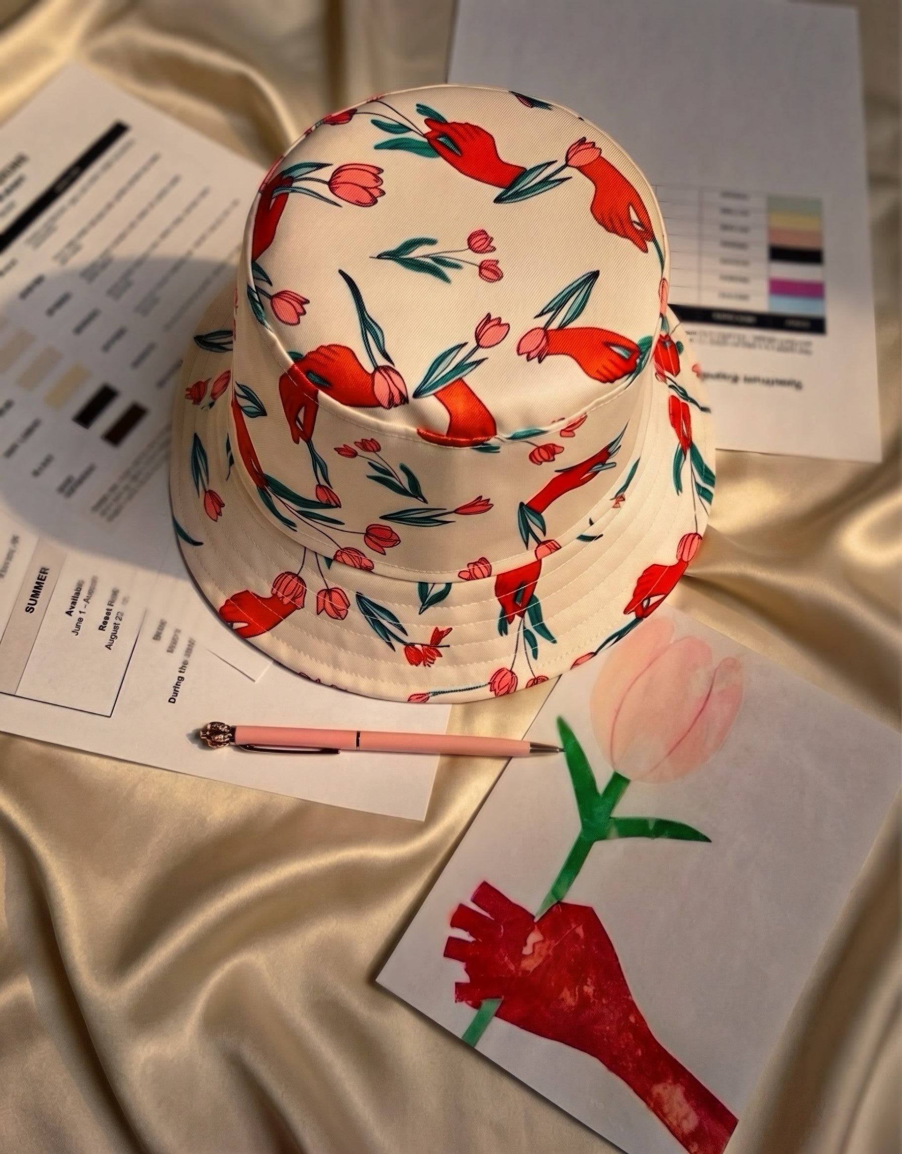 Image of Spectrum Expressions bucket hat featuring red hand and pink tulip artwork, styled with the original artwork and old and updated brand color swatches.