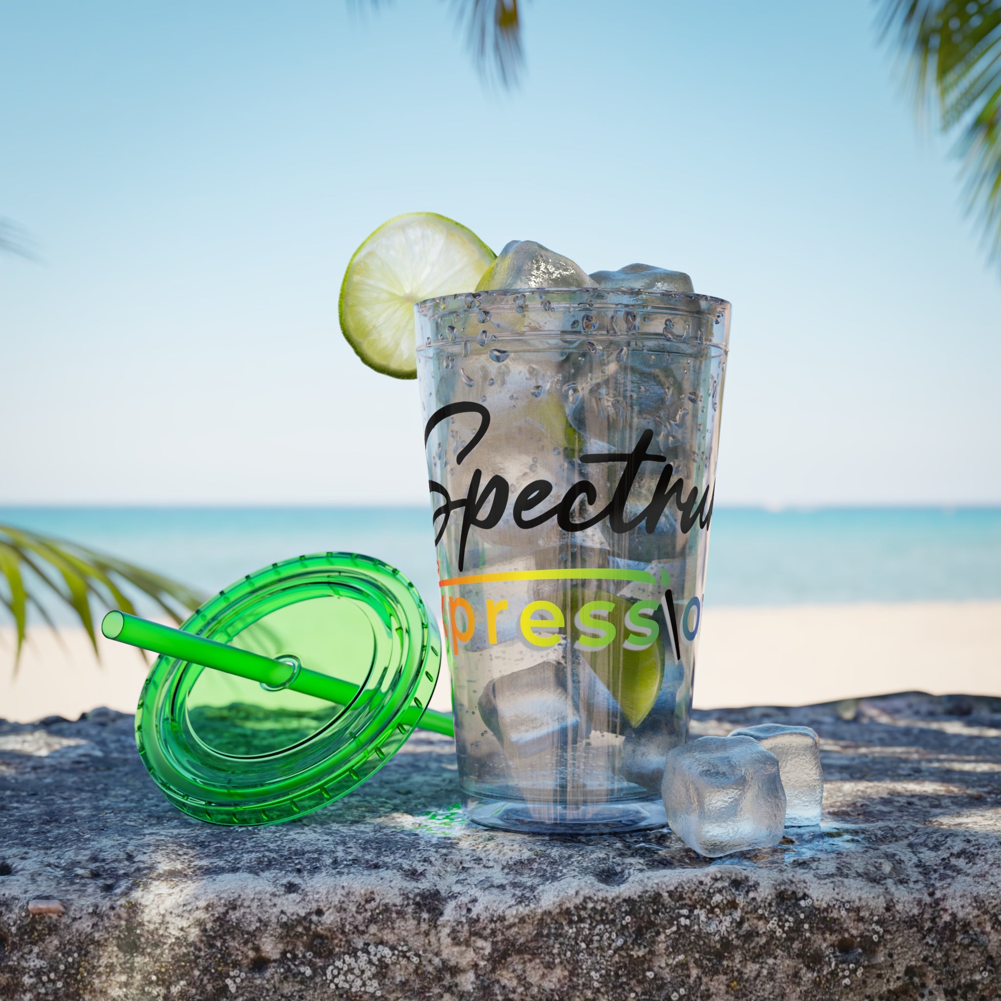 Image of A clear plastic tumbler with a green lid and straw on a stone surface at the beach, filled with ice and garnished with a lime slice. The cup has visible Spectrum Expressions branding on the front.