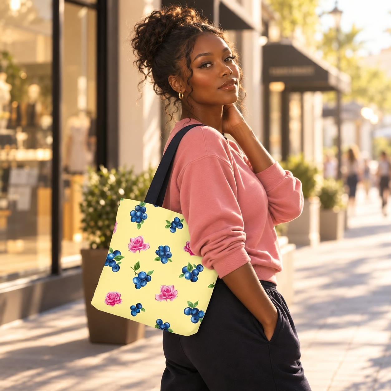 Image of A woman in a pink sweatshirt and dark pants carries a light yellow tote bag with a blue and pink floral print in a sunny shopping center.