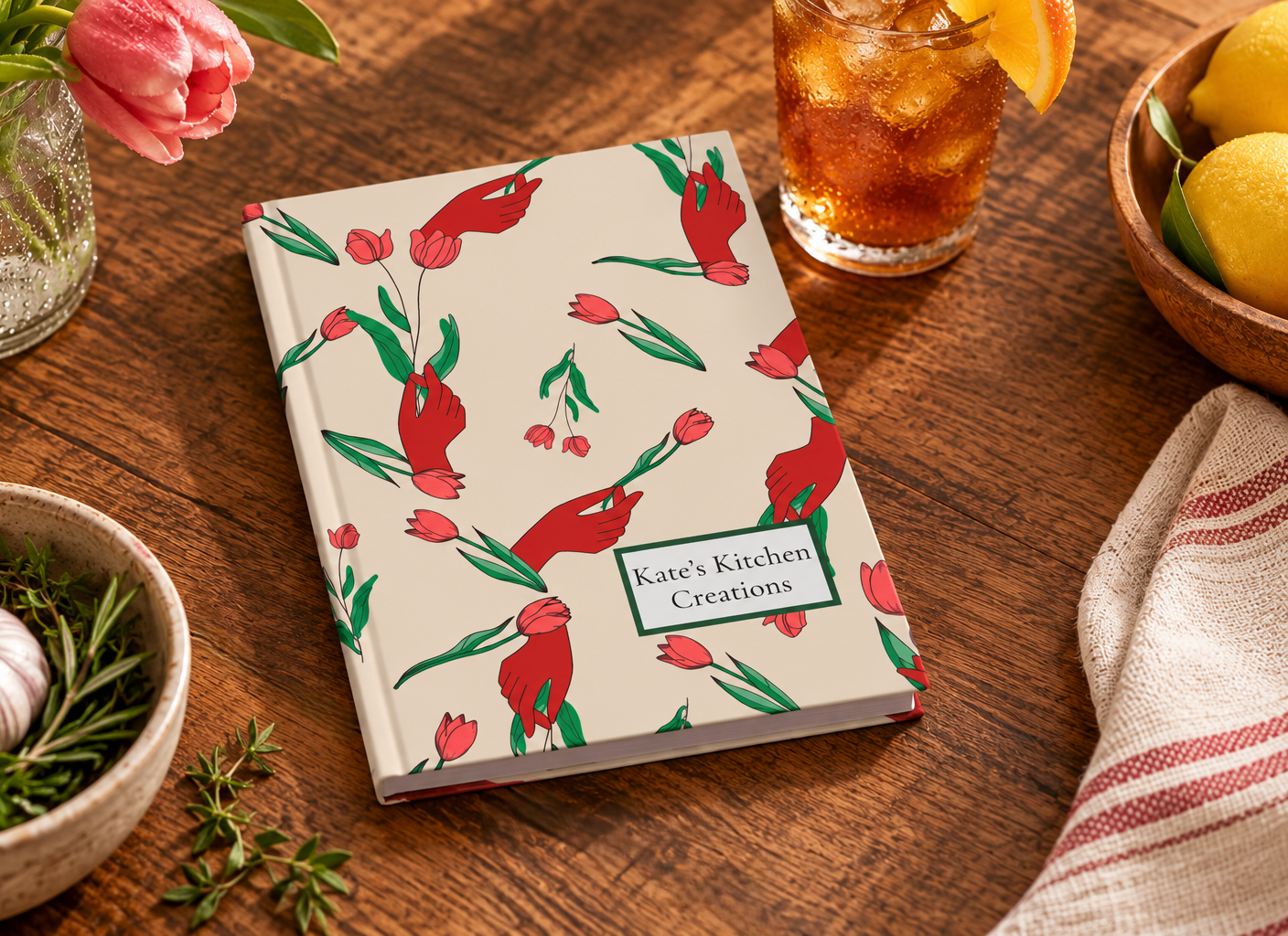 Image of A hardcover journal titled 'Kate's Kitchen Creations' with a cream cover decorated with red tulips and green leaves, styled on a wooden table with a drink, lemons, and herbs.