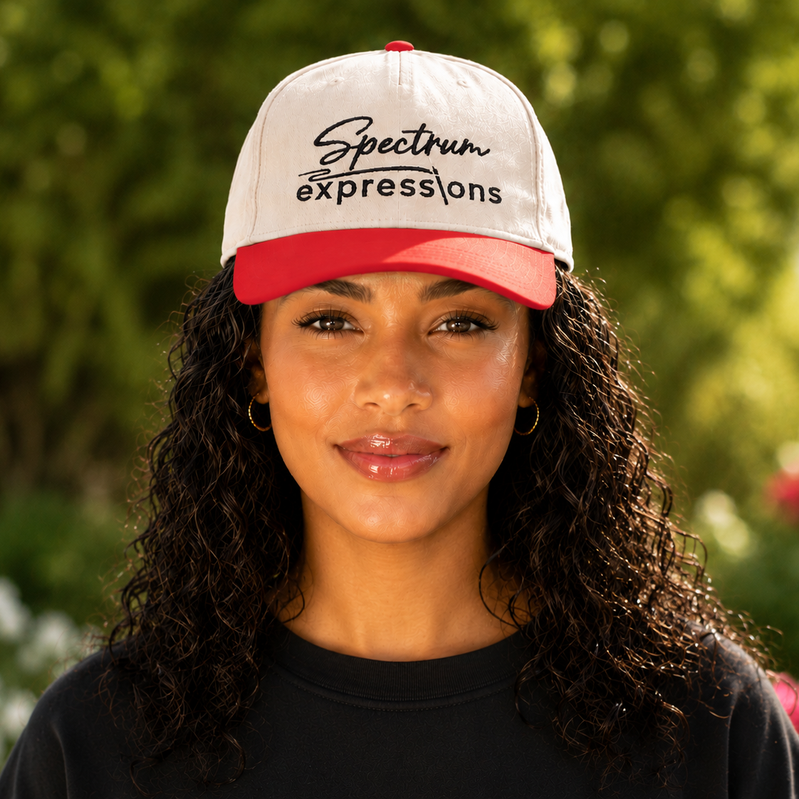 Image of A woman with curly dark hair wearing a white and red baseball cap with 'Spectrum expressions' printed on the front, photographed outdoors against a blurred green garden background.